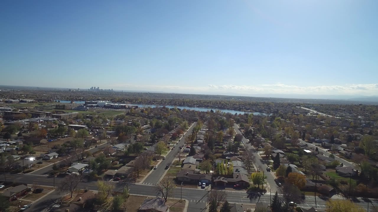 Opening aerial frame over Hidden Lake, Colorado, with neighborhood roofs and streets below, a blue lake beyond the trees, a distant skyline on the left horizon, and no visible people.