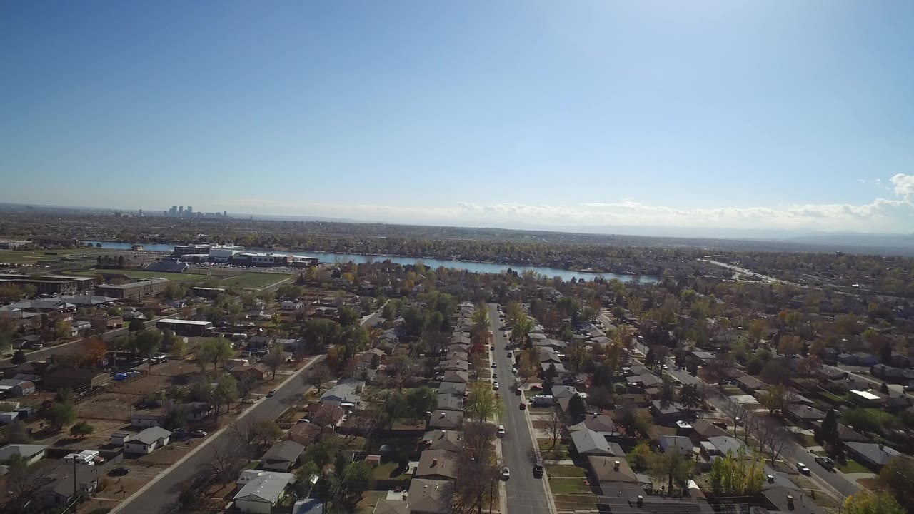 Aerial view of suburban houses, straight streets, trees, lake, and distant skyline.