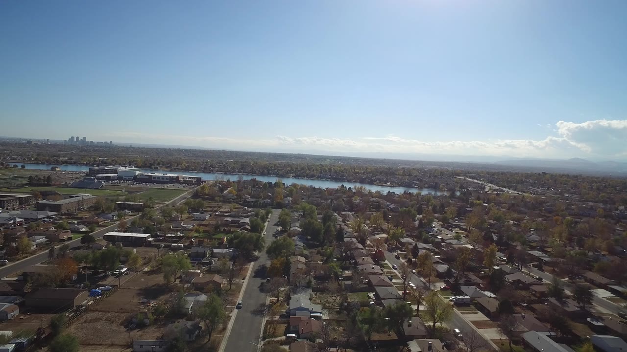 Aerial view over residential streets, homes, trees, and a distant lake under blue sky.