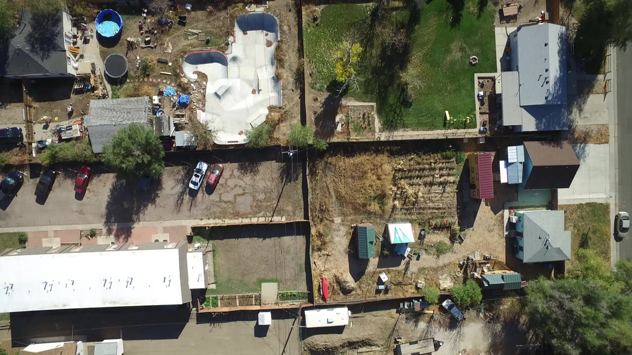 Aerial top view in Hidden Lake, Colorado with the white pool shell near center right, parked cars by a driveway, dry fenced lots, green lawn, trees, and no visible people.