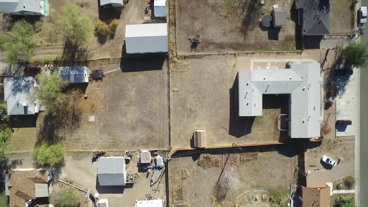 Overhead Hidden Lake, Colorado frame with a gray-roofed house, surrounding dry lots, sheds, trees casting shadows, parked vehicles, and very small visible people near property edges.
