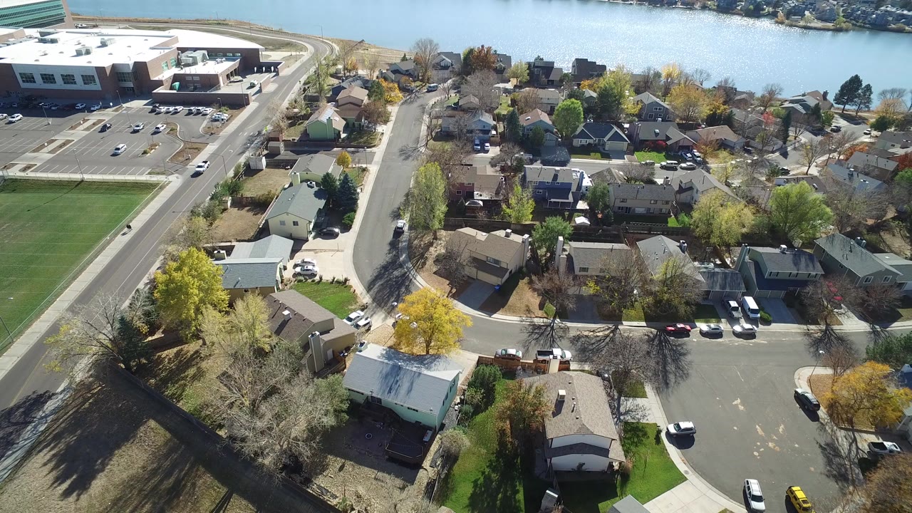 High aerial view in Hidden Lake, Colorado over residential rooftops, a curved street, lawns, parked cars, a grass field, and a waterway in the distance, with no visible people.