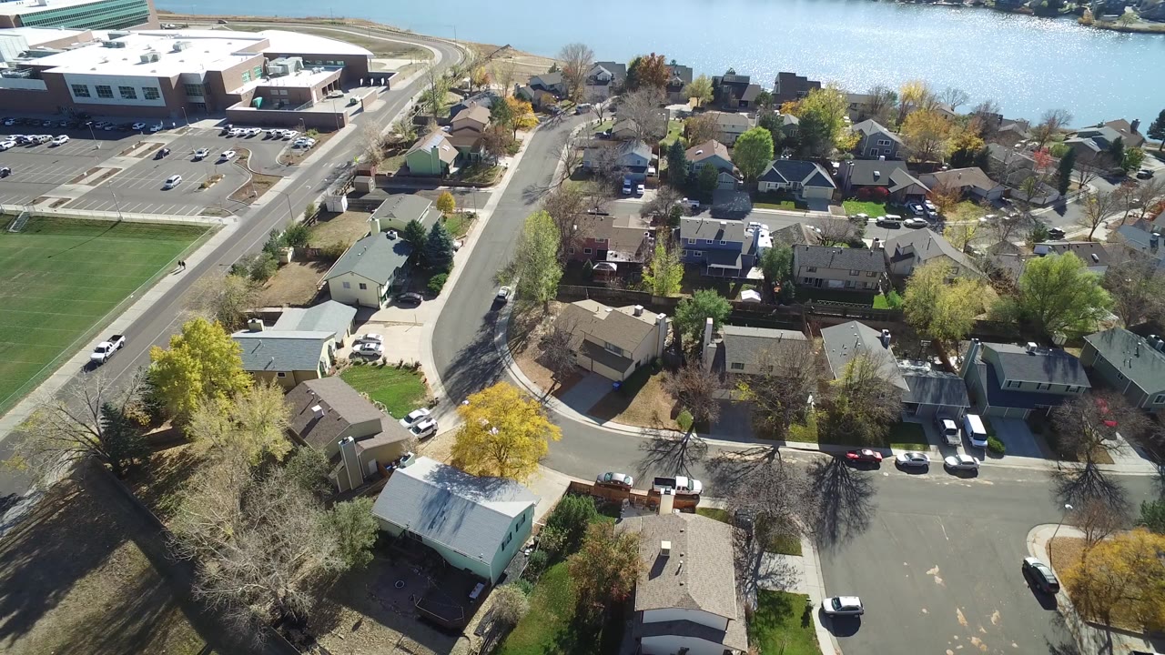 High aerial frame over Hidden Lake, Colorado homes and curved residential roads, with a large building, parking lot, green field, water in the background, and no visible people.