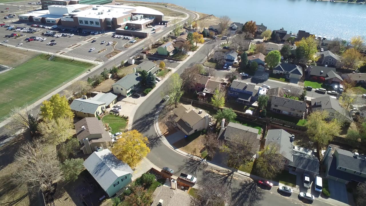 Aerial frame over Hidden Lake, Colorado showing residential rooftops, curved streets, green athletic fields, a large light-roofed building near the shore, blue lake water, autumn trees, and no visible people.