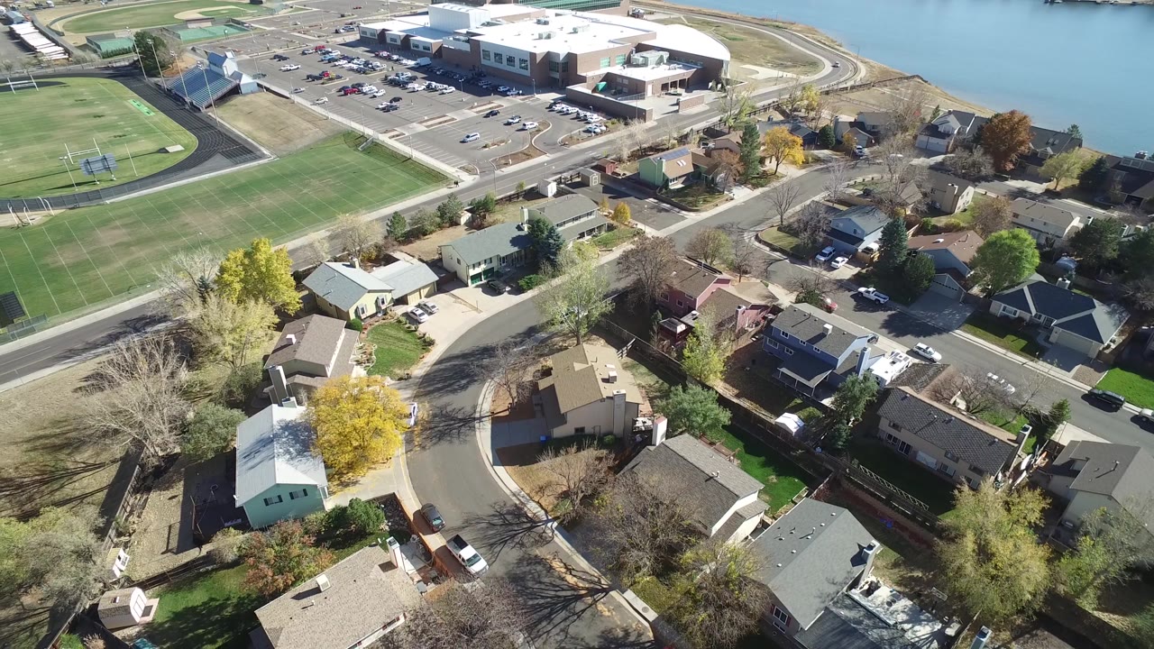Aerial neighborhood view with houses, trees, field, road, and lake edge.