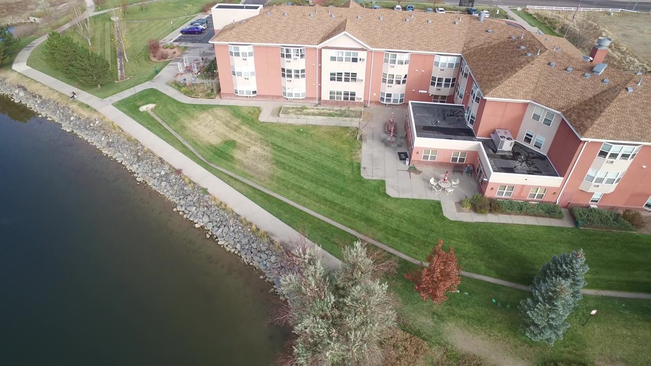 High-angle view of a lakeside residential building, lawns, paths, trees, and shoreline rocks.