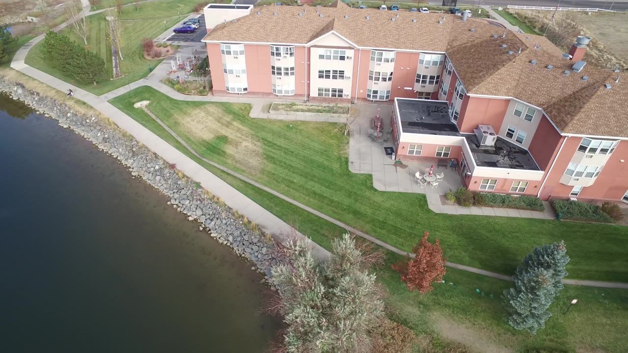 Early aerial frame in Hidden Lake, Colorado, with lake water at left, a rock shoreline, mowed lawns, intersecting paths, trees, parked cars, and the residential building filling the upper right, with no visible people in this frame.