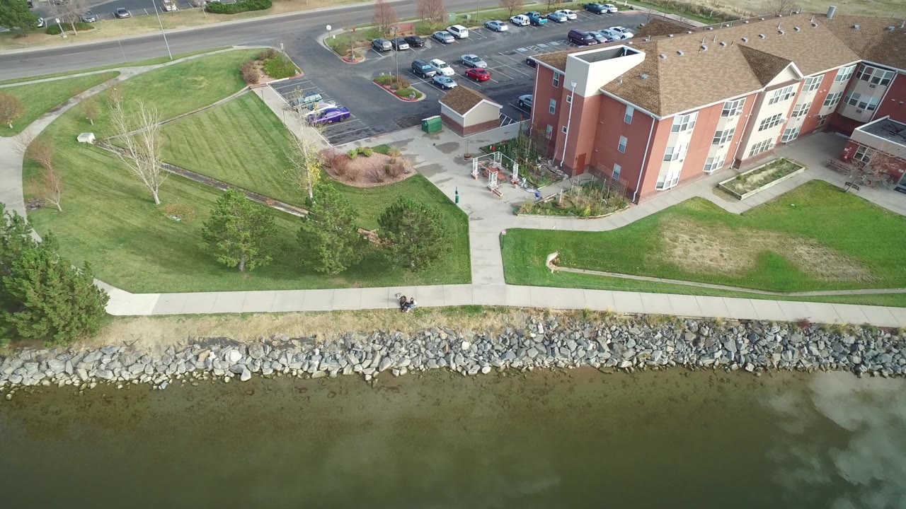 Aerial view of lakeside apartments, parking lot, lawns, sidewalks, and rocky shoreline.