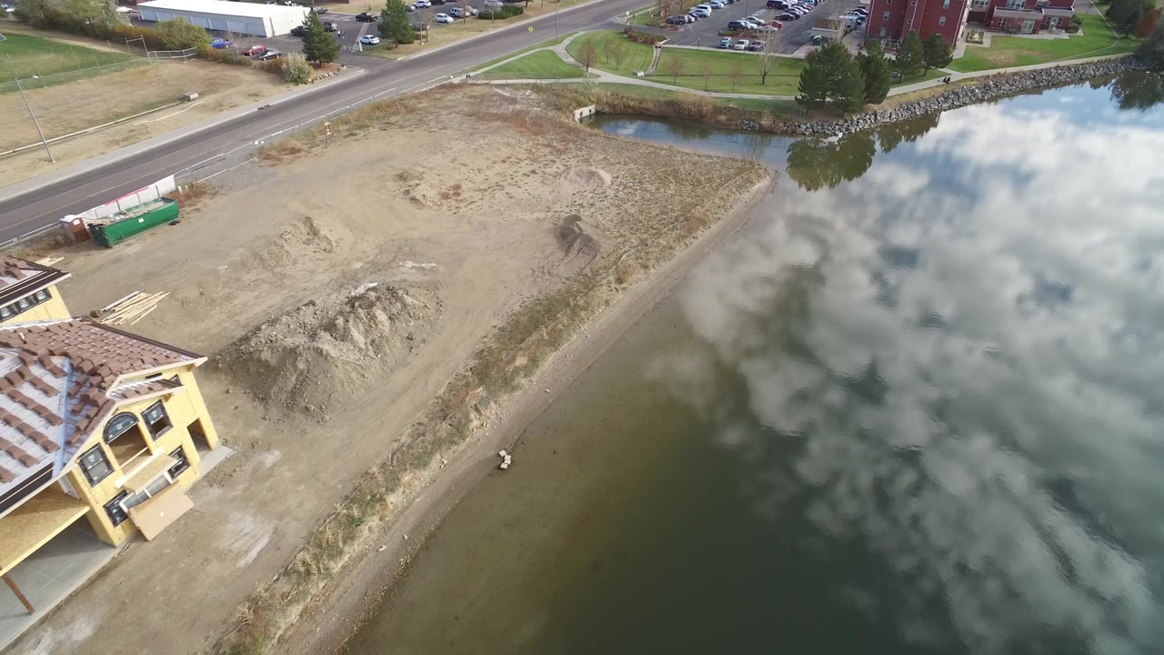 Bird's-eye frame over Hidden Lake, Colorado showing more of the yellow house roof, exposed dirt around the building, the curved lake edge, reflected clouds, roadside pavement, and no visible people.