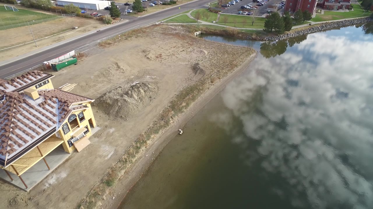Aerial pass over a house roof beside lake water and a dirt construction lot.