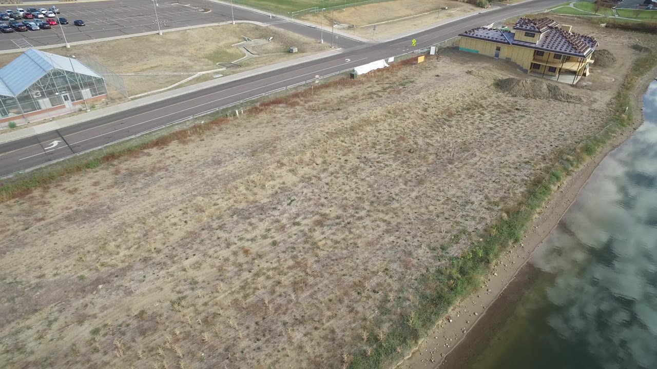 Hidden Lake, Colorado aerial frame looking across dry graded ground toward a paved road, a greenhouse-like structure, a partly framed building, and parked vehicles, no visible people.