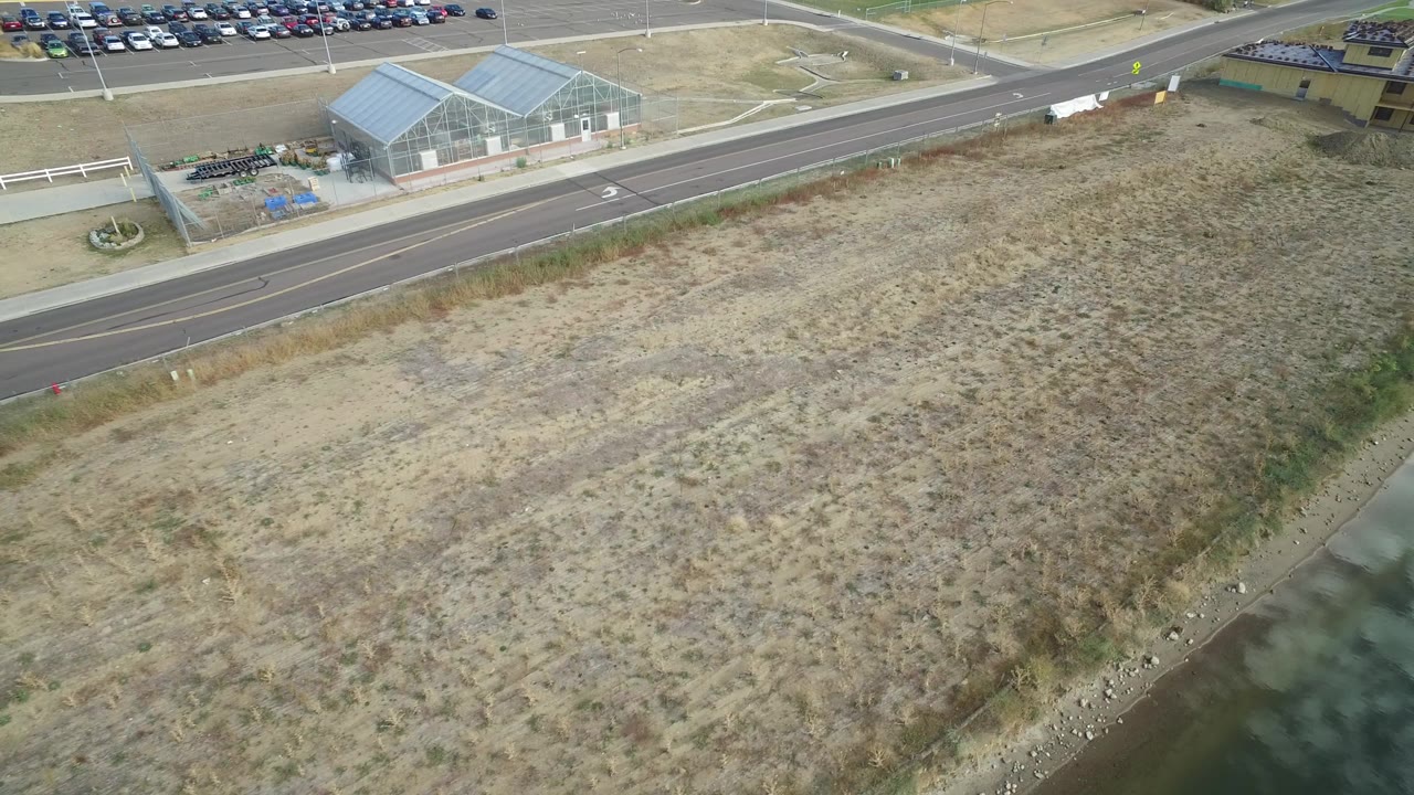 Aerial frame over Hidden Lake, Colorado showing a dry grassy field, roadside, greenhouse-like industrial building, parked cars, and no visible people.