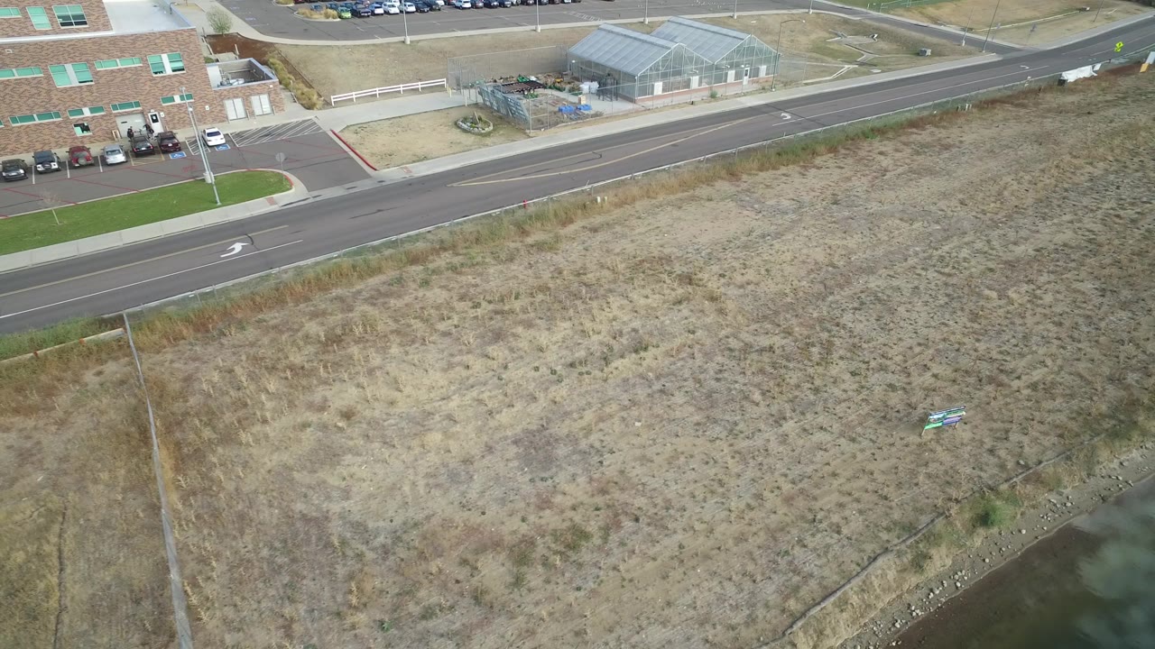 High oblique aerial view in Hidden Lake, Colorado showing dry grassland, a small water channel, a paved road, parked cars, brick buildings, and no visible people.