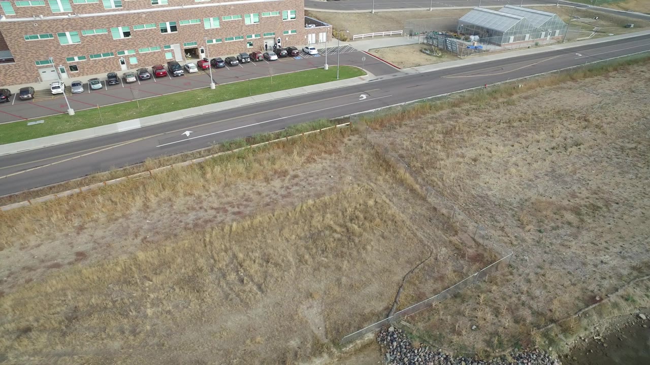 High aerial frame in Hidden Lake, Colorado showing a paved road beside a brick building and parked cars, with dry open ground below and no visible people.