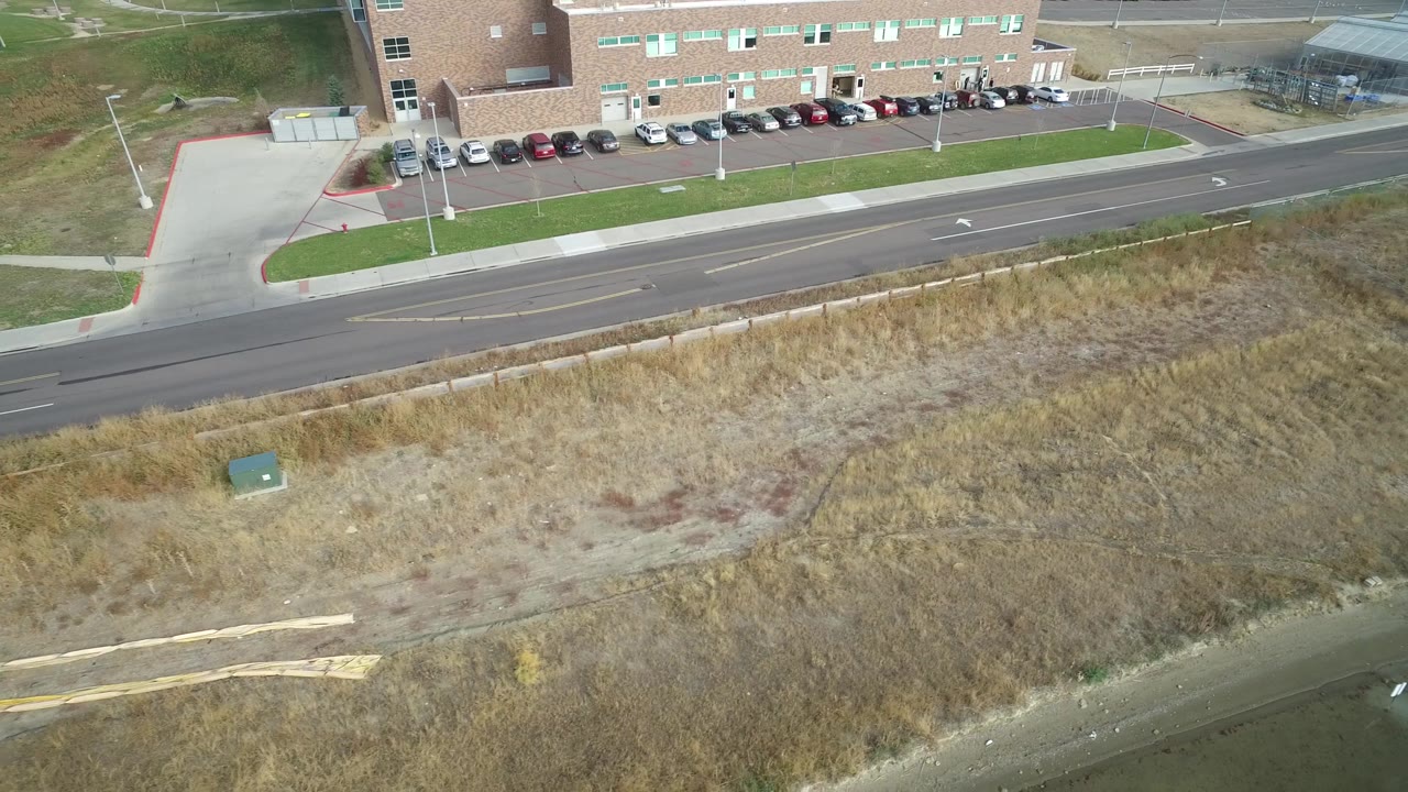Aerial view in Hidden Lake, Colorado with the road still mostly empty, dry scrubby grass below, a brick building and parking row above, and no visible people.