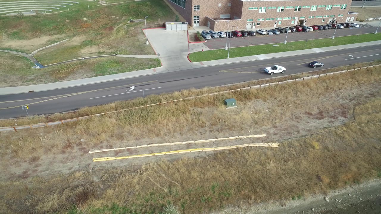 Hidden Lake, Colorado aerial view with two vehicles on the road, dry grass in the foreground, and parked cars near a brick building, with no visible people.