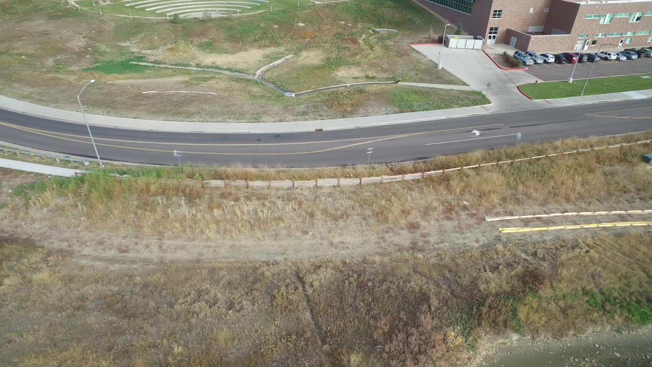High aerial view of road, sidewalk, dry grass, and a building corner near water.