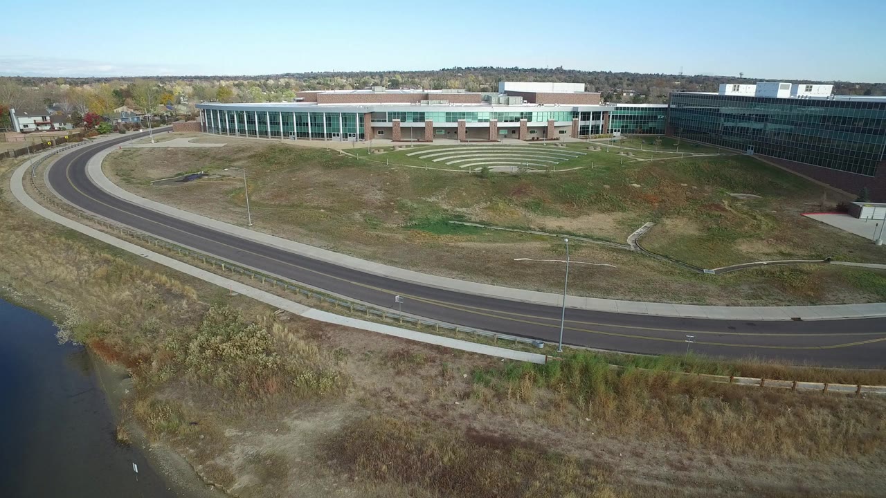 Opening aerial frame in Hidden Lake, Colorado showing a curved road, water at lower left, dry grass, and a modern educational facility under clear daylight with no visible people.