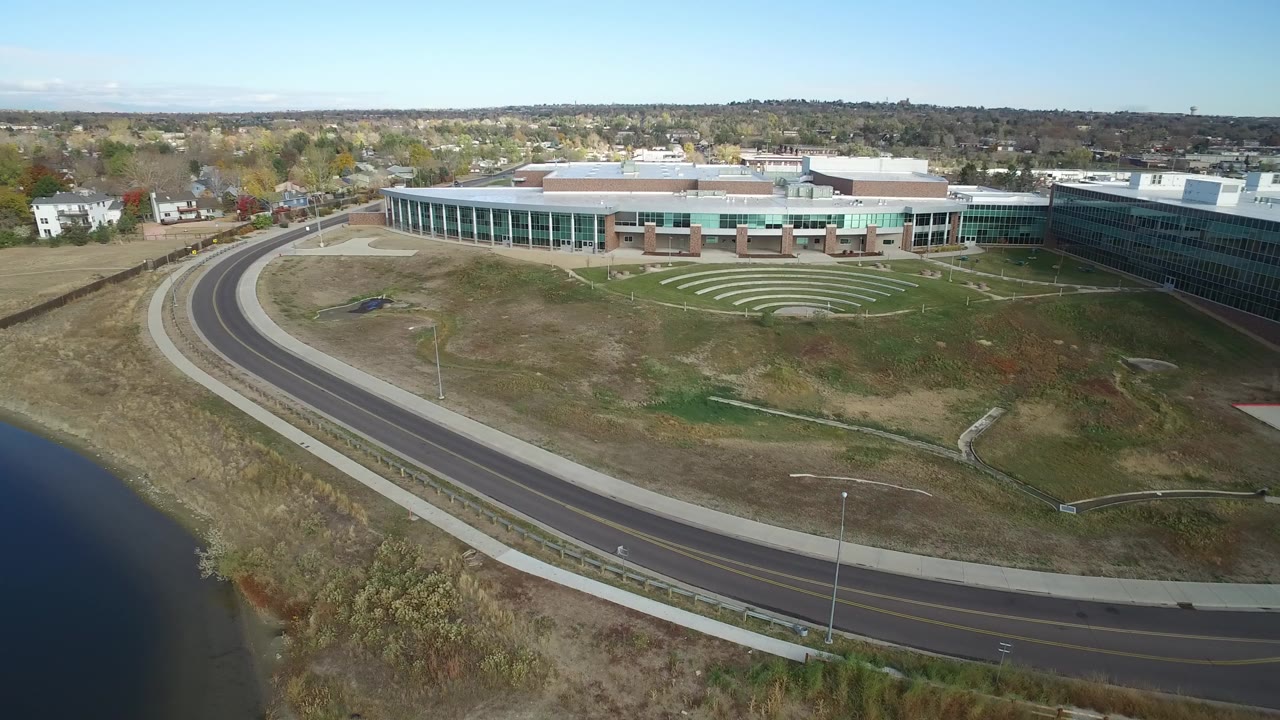 At 0:00, an elevated view over Hidden Lake, Colorado shows dark water, a curved road, dry grass, a large glass-fronted building, distant homes, and no visible people.