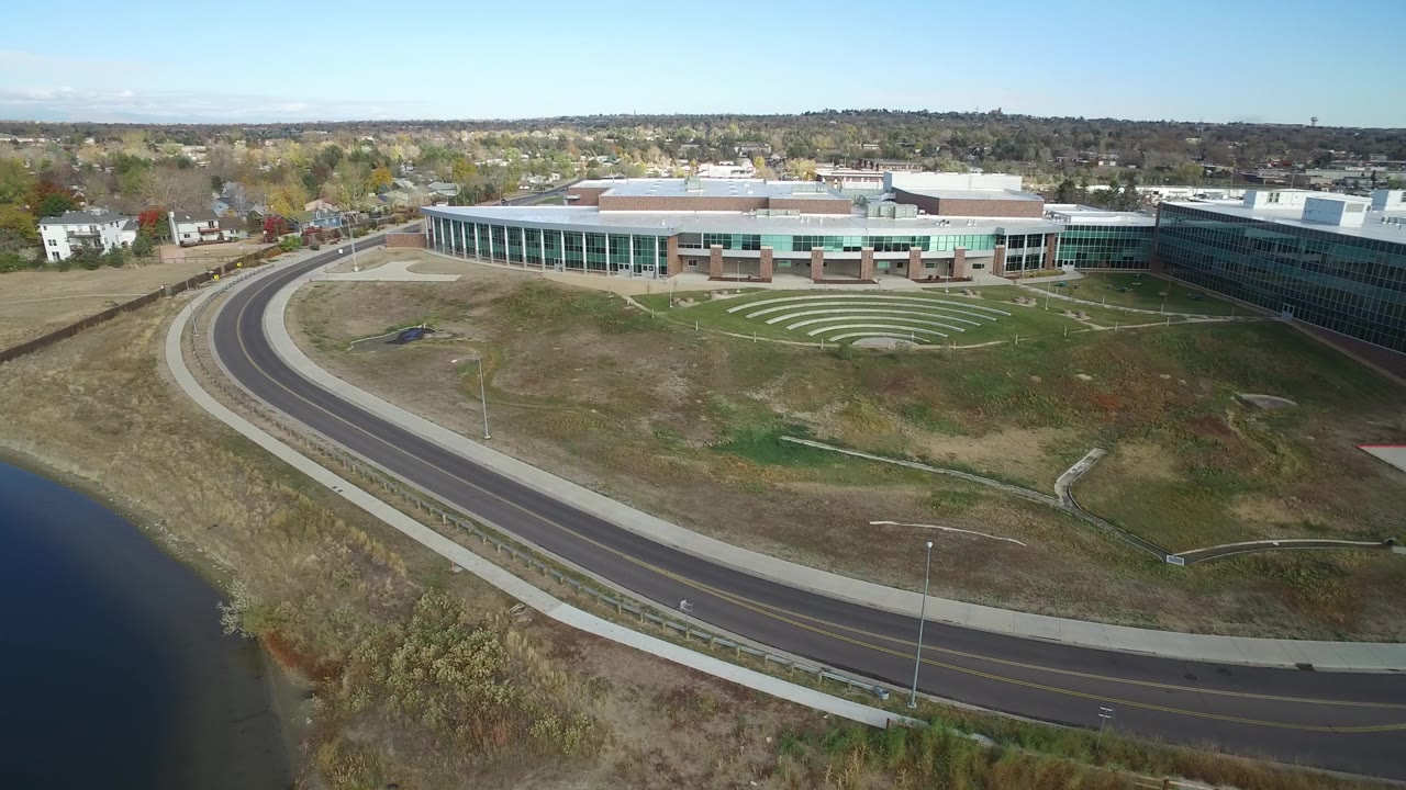 At 0:01, the drone perspective keeps the lake at lower left, the curving road through the center, a broad modern building beyond the grass, and no visible people.