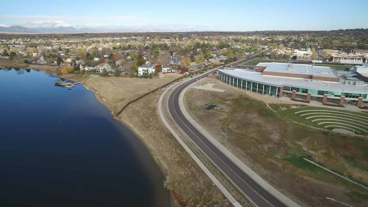 High aerial frame over Hidden Lake, Colorado showing dark lake water at left, a dry shoreline, a curved road, a glass-fronted building, distant homes, hills on the horizon, and no visible people.