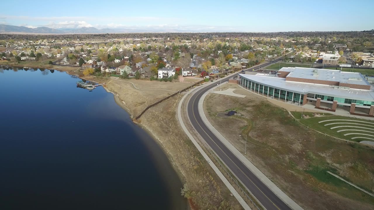 Wide aerial of lake, arid shore, curved road, building edge, and homes.