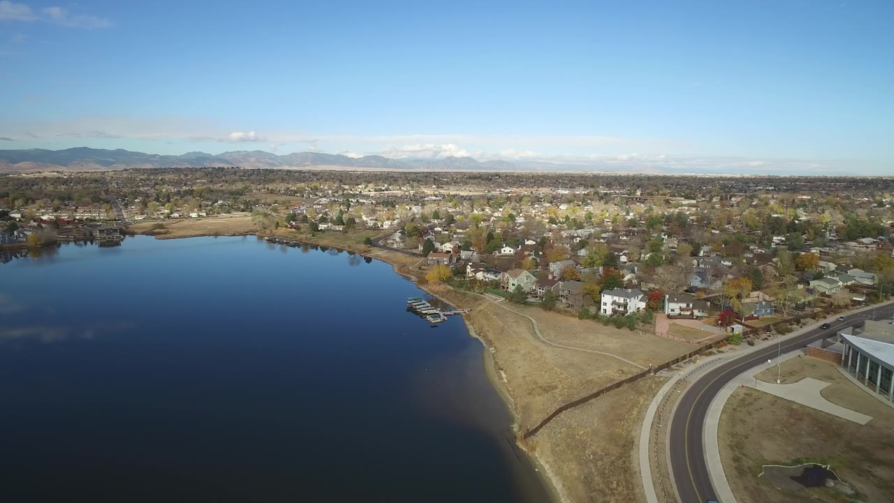 Aerial view over Hidden Lake, Colorado showing dark blue water on the left, a dry shoreline, lakeside homes, a curving road, distant mountains, and no visible people.