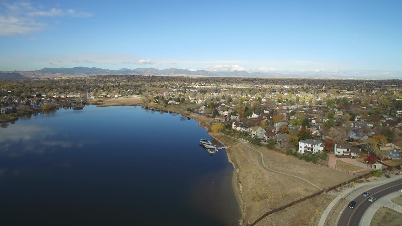 Opening aerial frame over Hidden Lake, Colorado, with dark-blue water in the foreground, a curved residential shoreline, distant mountains, and no visible people.