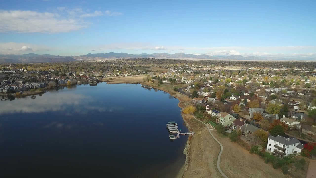 Aerial view over Hidden Lake, Colorado, with dark blue water, reflected clouds, shoreline homes, a small dock, distant mountains, and no visible people.