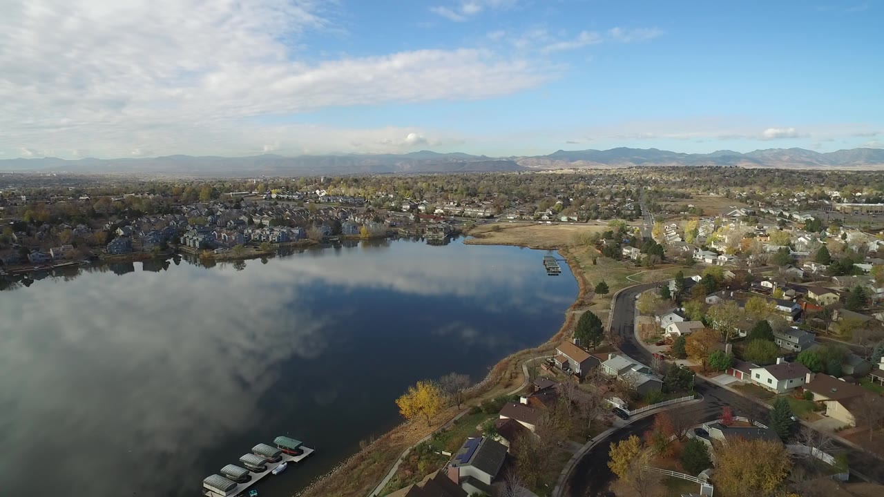 Opening aerial frame over Hidden Lake, Colorado, with reflective water in the foreground, homes along the shore, distant mountains under patchy clouds, and no visible people.