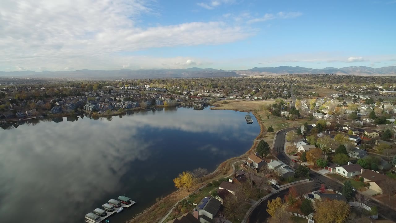 Wide aerial lake view with houses along the shore and clouds reflected in water.