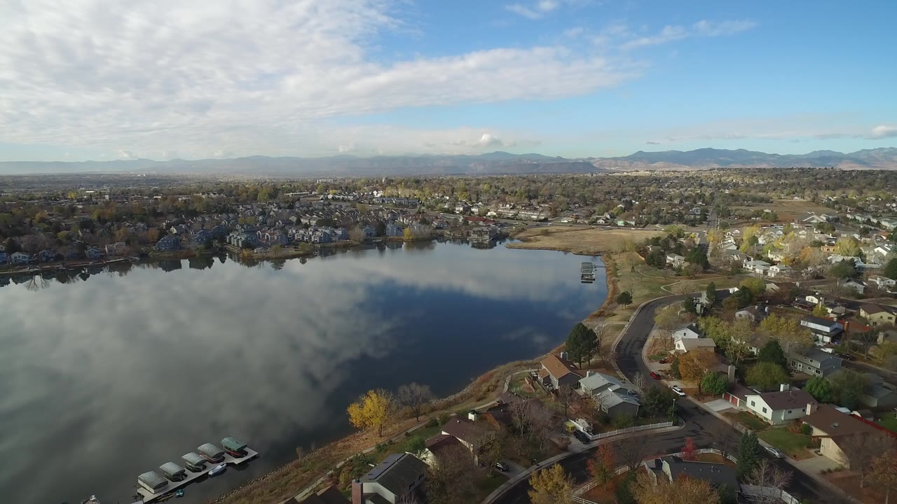 Aerial view over a reflective lake beside shoreline homes and distant mountains.
