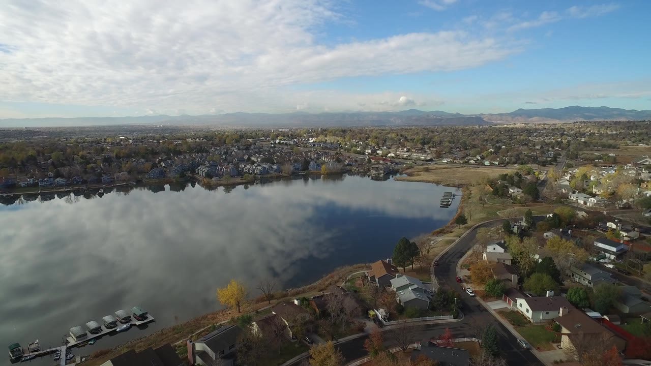 Opening aerial frame over Hidden Lake, Colorado, with reflective water in the foreground, shoreline homes and roads to the right, distant hills on the horizon, and no visible people.