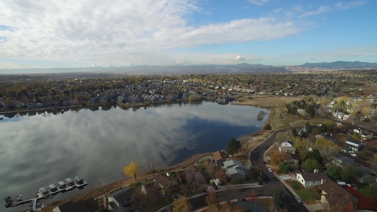 High aerial view of houses, roads, trees, and a lake under broken clouds.