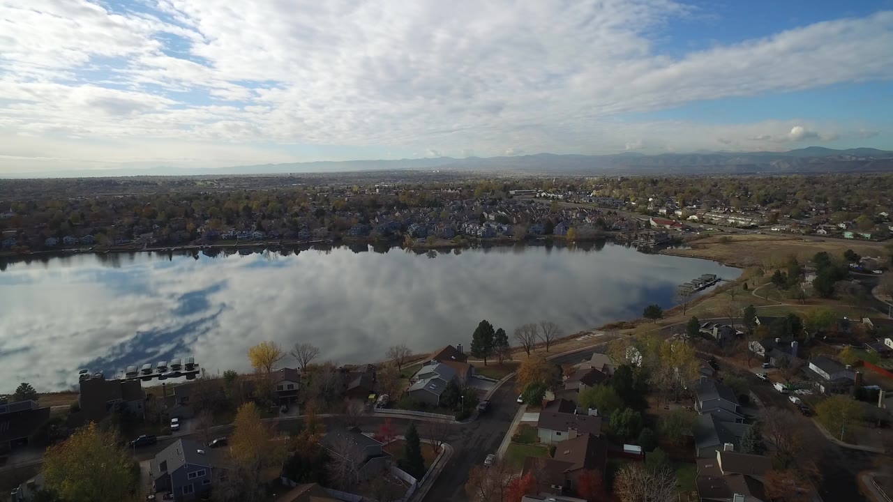 Aerial frame over Hidden Lake, Colorado showing cloud reflections on the water, houses and roads in the lower edge, tree cover beyond the lake, and no visible people.