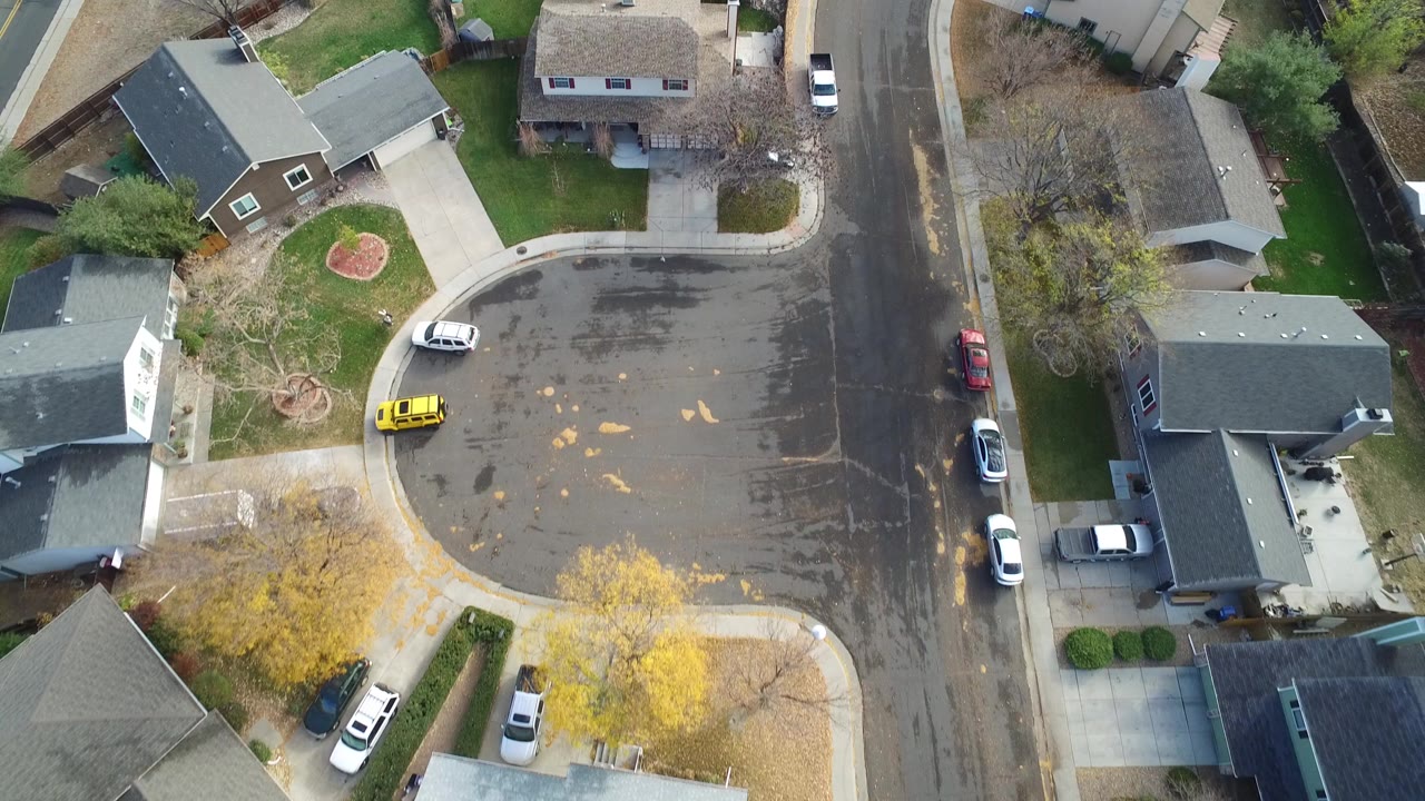 Aerial frame over Hidden Lake, Colorado showing a curved residential street, detached homes, driveways, a yellow car, white cars, and no visible people.