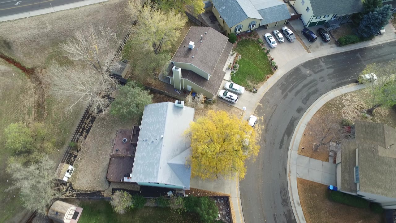 Overhead frame from Hidden Lake, Colorado showing detached homes around a curved residential street, dry yards, yellow and leafless trees, parked cars, and no visible people.