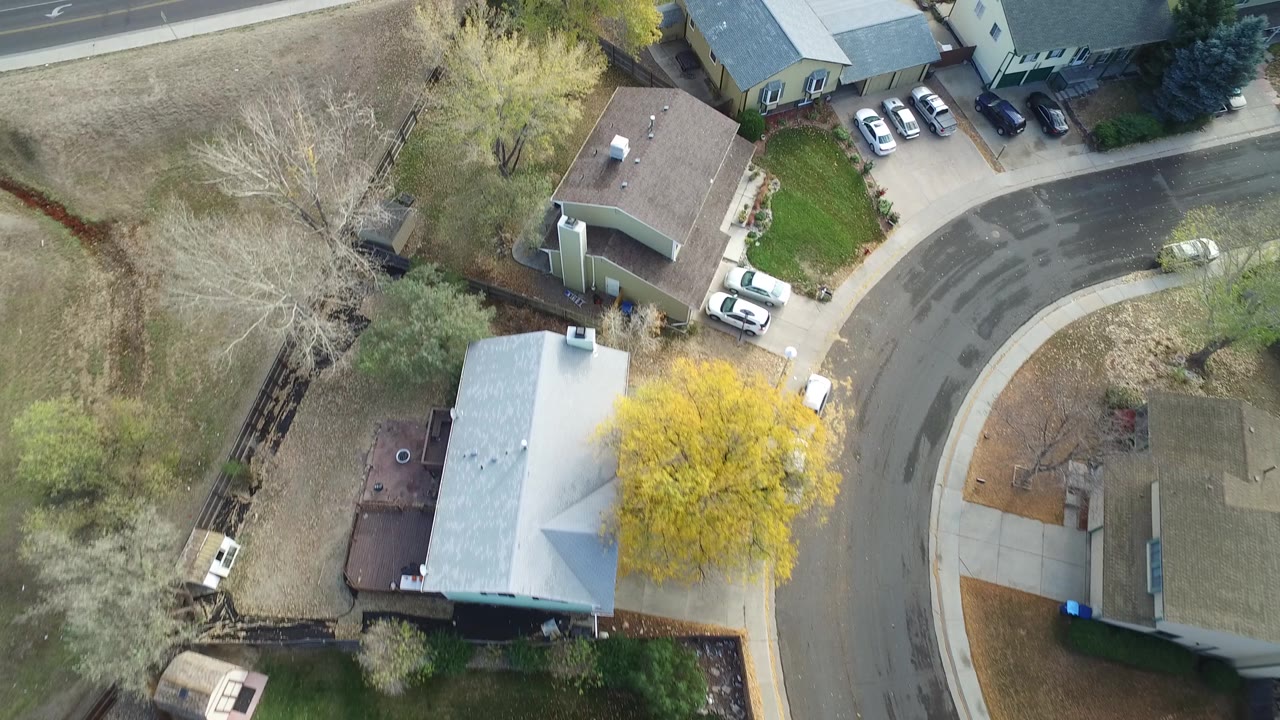 Aerial frame from Hidden Lake, Colorado with a brown-roofed house near center, a light-roofed house below, open grass, trees, parked cars, and no visible people.