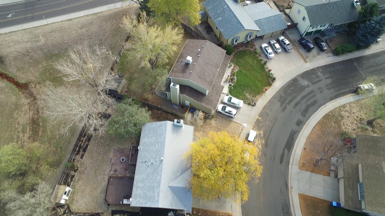 Top-down neighborhood view with houses, a road, yards, trees, and parked vehicles.