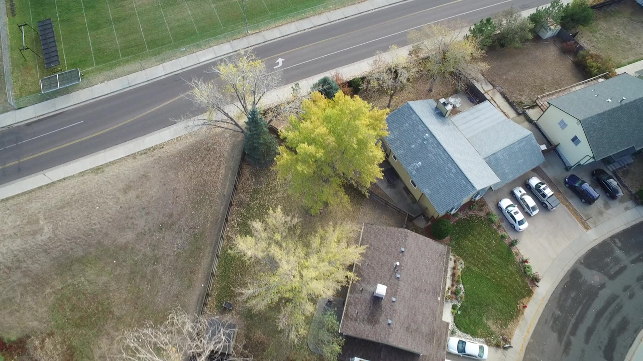 Overhead view of a road, homes, parked cars, trees, and open ground beside a sports field.