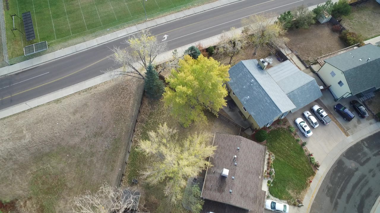 A later overhead view in Hidden Lake Colorado keeps the road and sports field across the top while detached houses, driveways, trees, and dry open lots fill the lower frame, with no visible people.