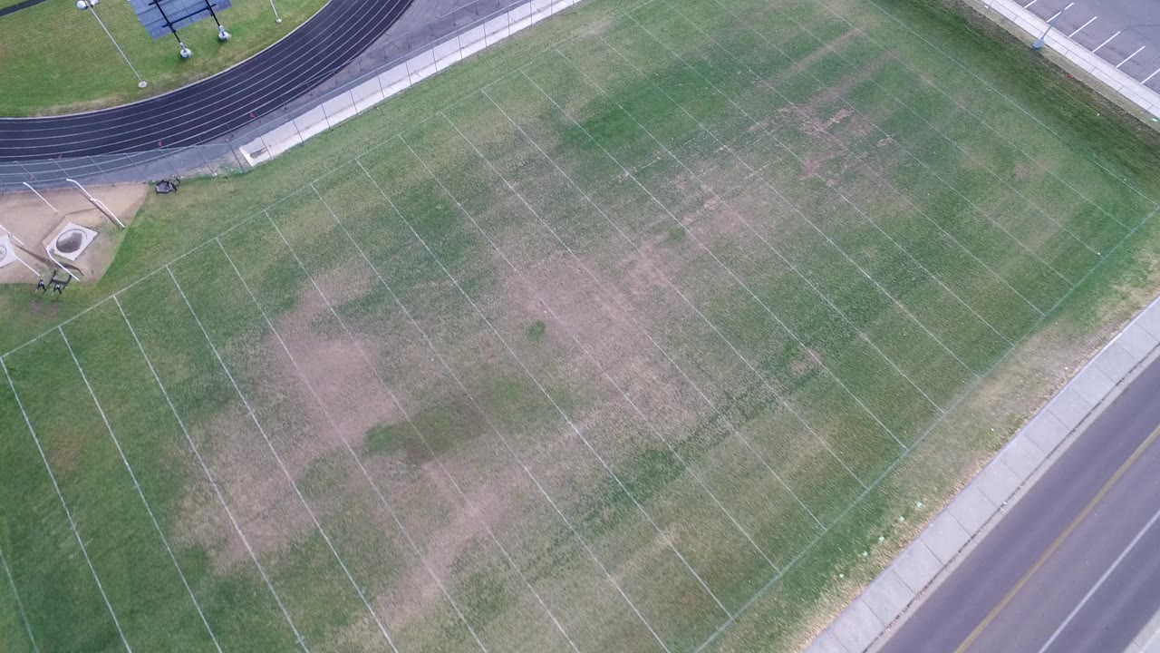 Overhead aerial frame in Hidden Lake, Colorado showing a green football field with white yard lines, worn turf patches, a curved black running track, and no visible people.