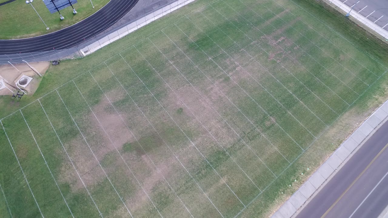 Drone-style overhead frame in Hidden Lake, Colorado showing the field centered more tightly, with pale yard markings, patchy grass, the track corner, and no visible people.