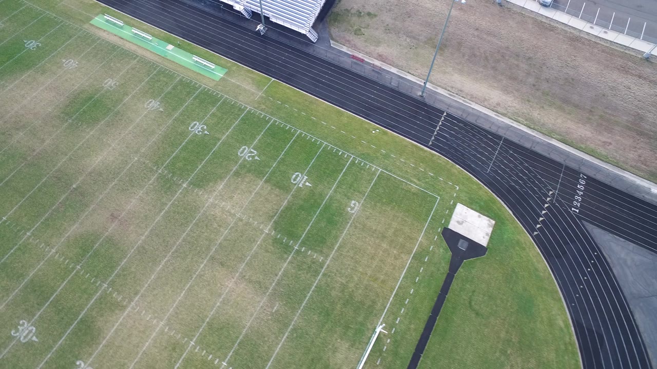 High aerial frame in Hidden Lake, Colorado with faded football yard markings crossing patchy turf, the running track curving along the right side, and no visible people.