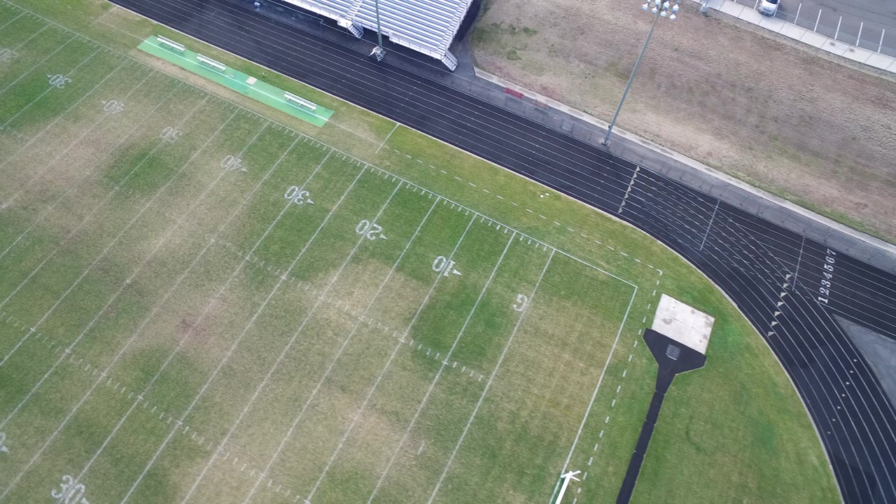 Drone view over a football field with parched turf, track lanes, and bleachers.