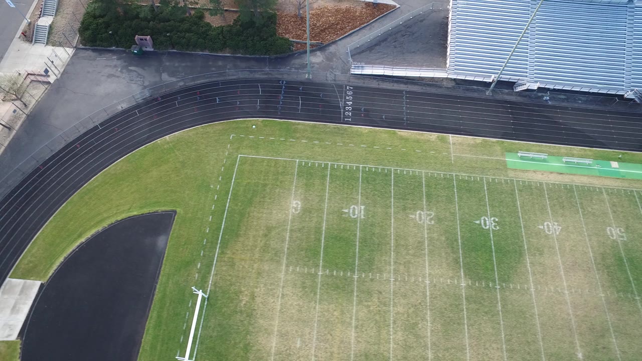 Overhead aerial view in Hidden Lake, Colorado showing the left side of a marked football field, a black curved running track, bleachers, road edges, trees, and no visible people.