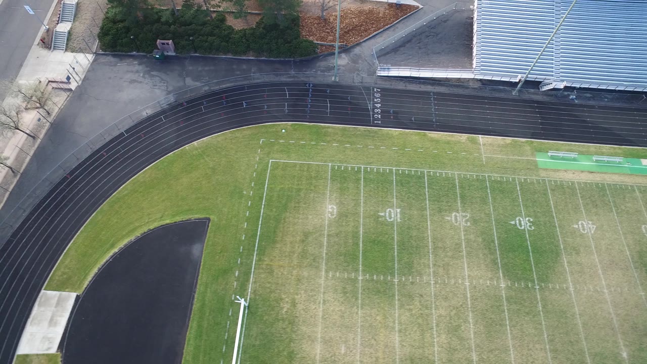 Top-down football field with track curve, bleachers, trees, and road.