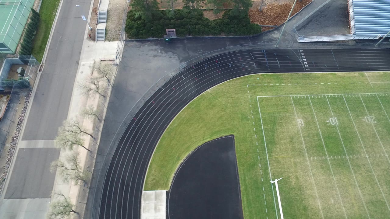 High aerial frame in Hidden Lake, Colorado showing a green football field bordered by a black running track, a road, and tennis courts, with no visible people.