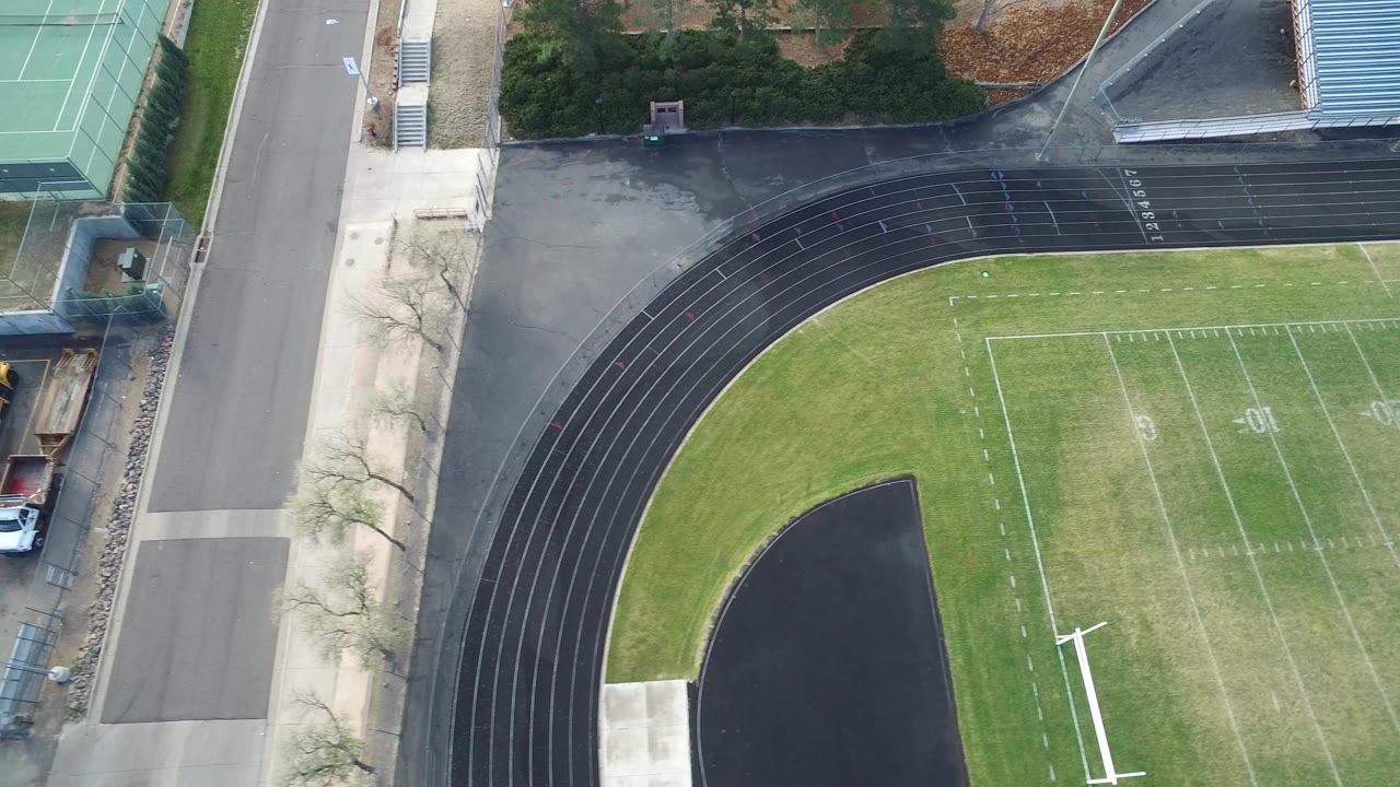 Top-down sports field and track with tennis courts, road, trees, and bleachers.