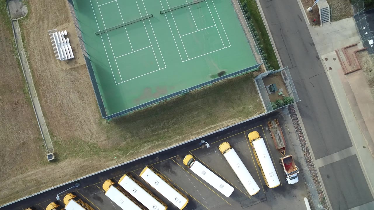 High aerial frame in Hidden Lake, Colorado with two green tennis courts at the top, dry grass through the center, a street on the right, parked school buses below, and no visible people.